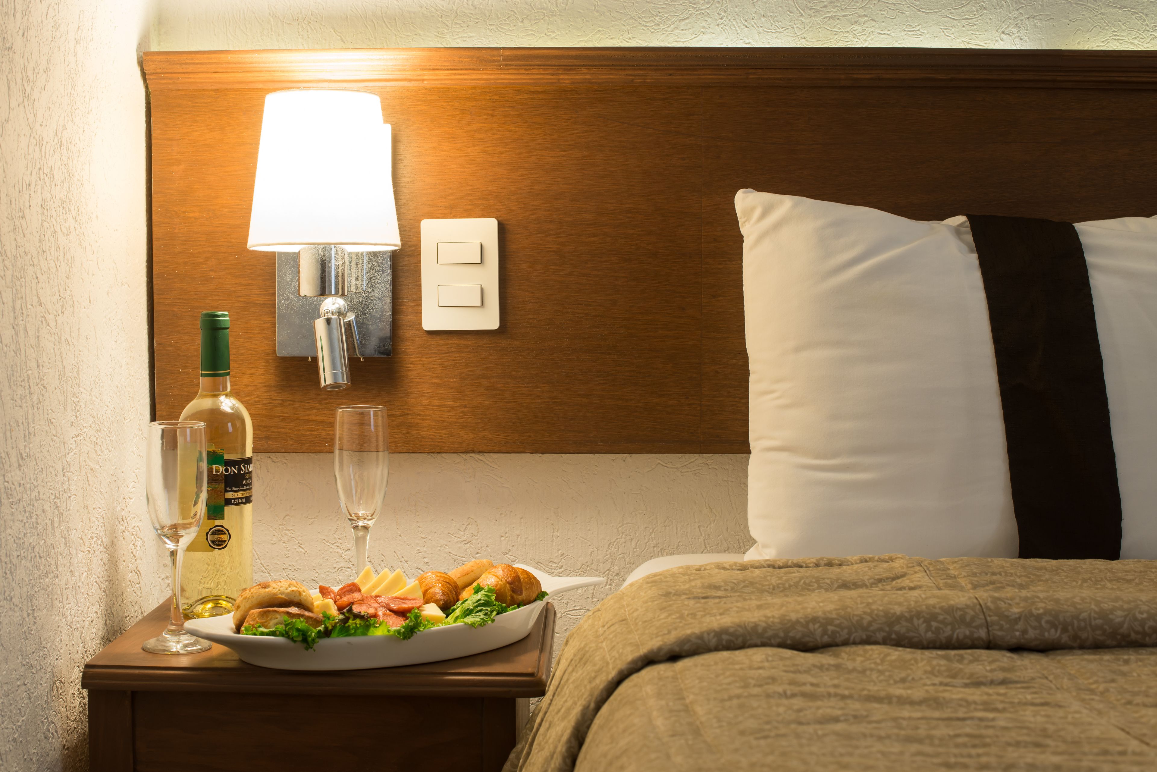 A neatly made bed with a white pillow and a brown blanket, a bedside table with a bottle of wine, two glasses, and a plate of food.