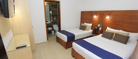 A hotel room with two beds, a wooden headboard, a bedside table with a lamp, a mirror, and a bathroom visible through an open door.