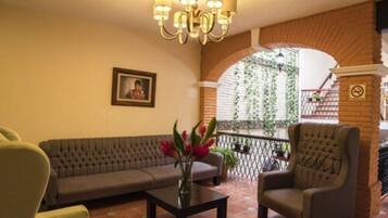 Un hall con un sofá, sillón y una mesa con flores. Hay una araña, un cuadro en la pared y un balcón con plantas.