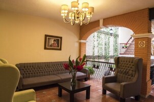 Un hall con un sofá, sillón y una mesa con flores. Hay una araña, un cuadro en la pared y un balcón con plantas.