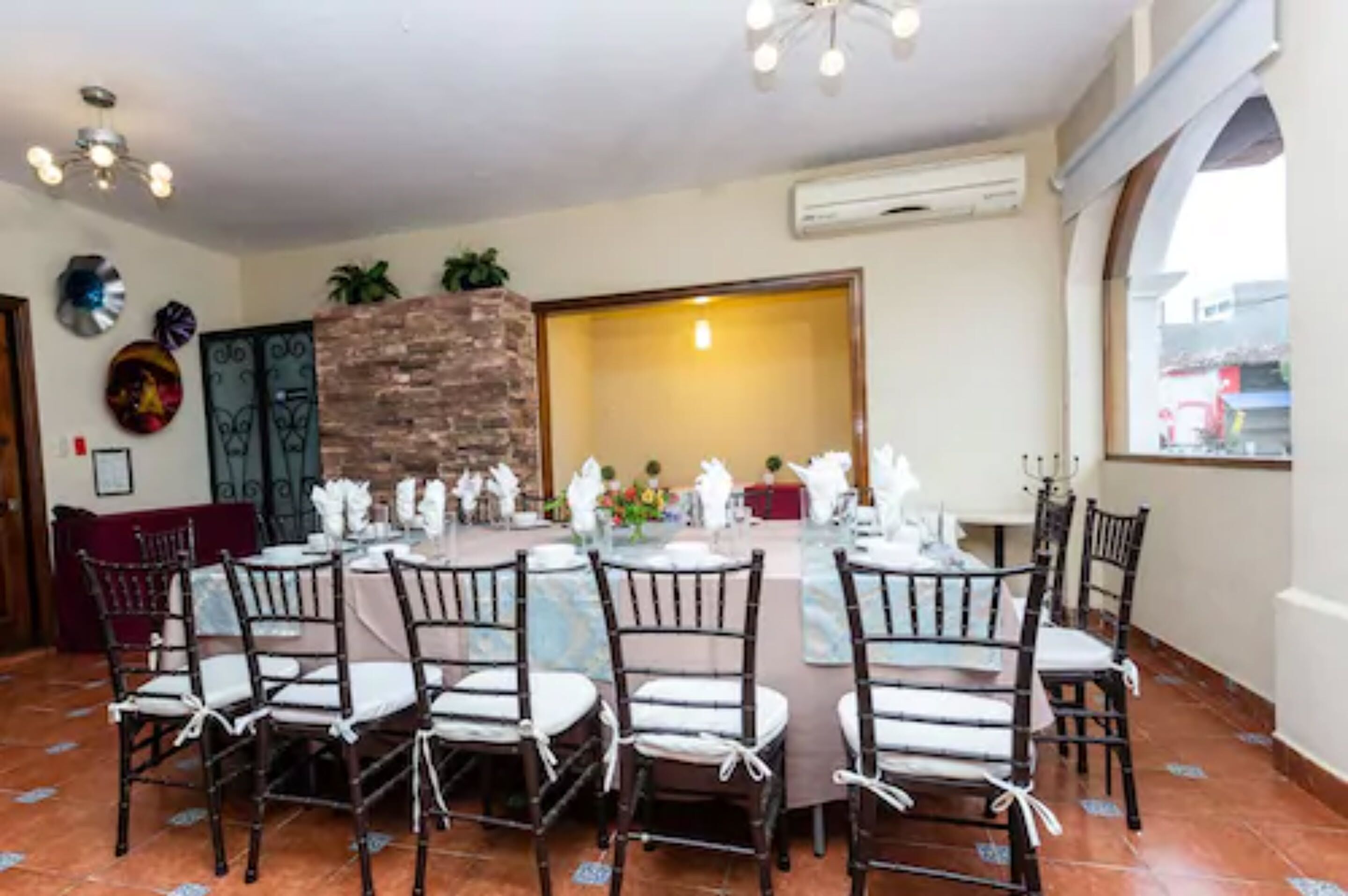 A dining area with a long table set for six, surrounded by chairs. There is a stone wall and a door leading to another room.