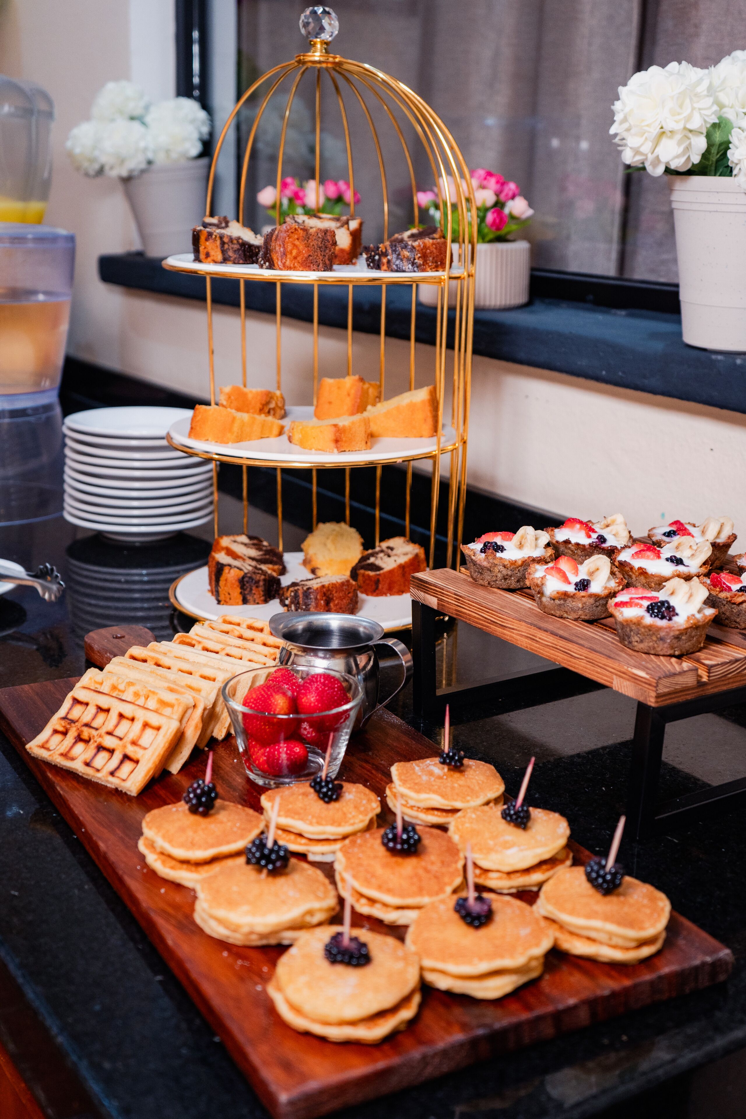 Uma mesa de buffet com diversos doces e pastelaria, incluindo waffles, panquecas e um suporte escalonado para bolos com vários produtos de pastelaria.