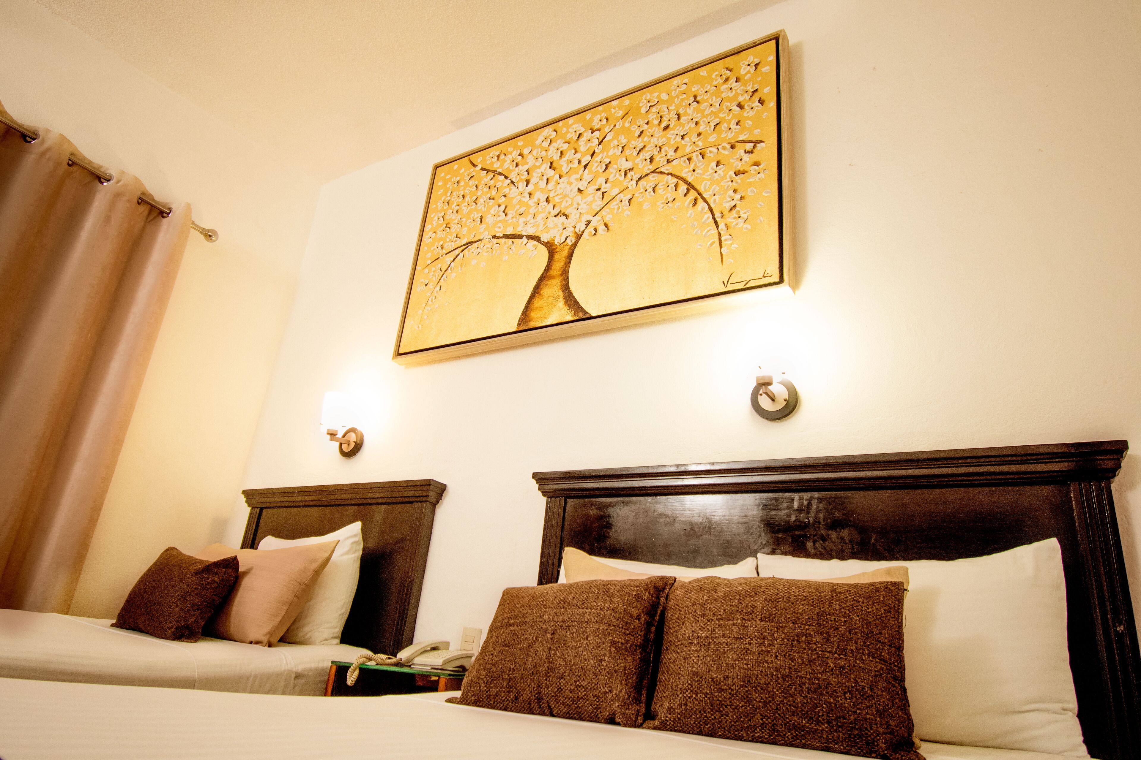 A hotel room with two beds, a painting of a tree, and a nightstand with a phone.