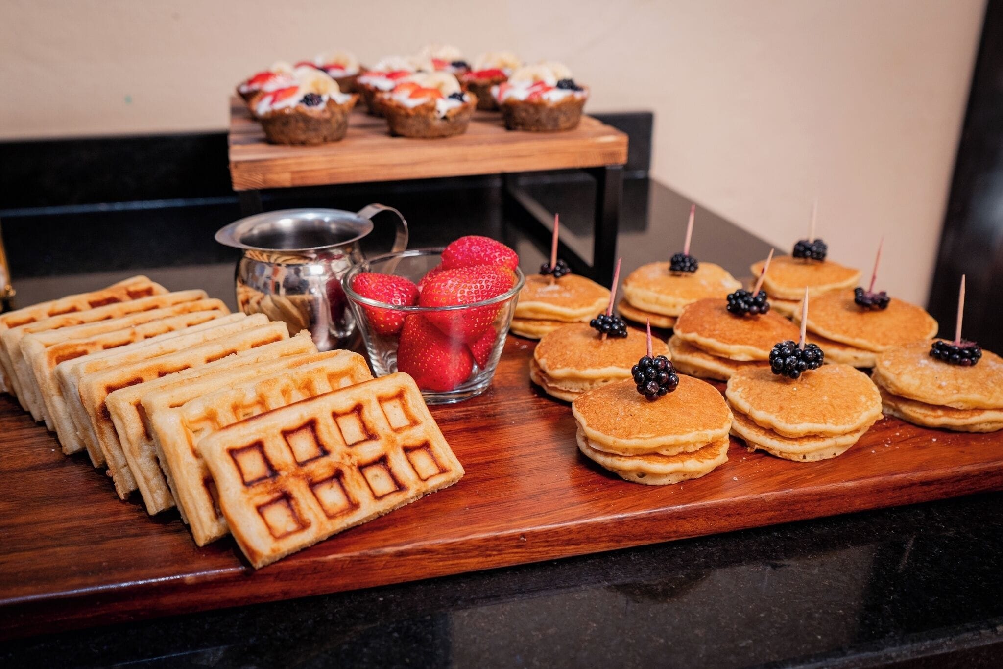 Verschiedene Frühstücksleckereien, darunter Waffeln, Pfannkuchen und Früchte-Torten, auf einer Holzservierplatte.