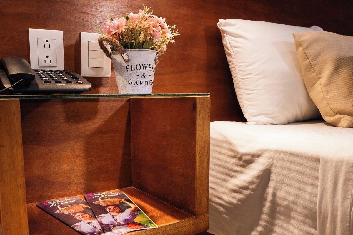 Una mesita de noche de madera con un jarrón de flor blanca, un teléfono negro y una revista.