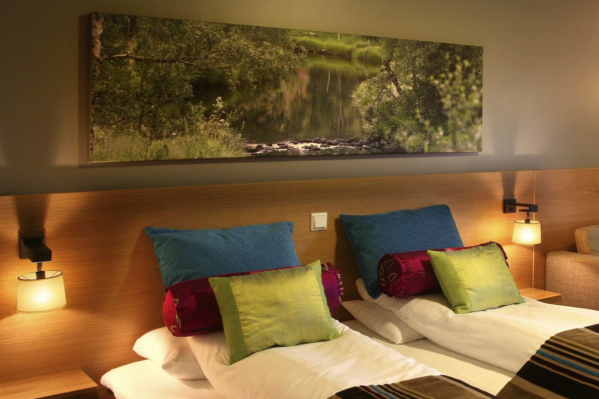 A hotel room with a large bed, two pillows, a nightstand with a lamp, and a painting of a forest scene on the wall.
