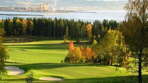 A golf course with bunkers and a view of a body of water and industrial structures in the background. - Scandic Stiklestad (Verdal)