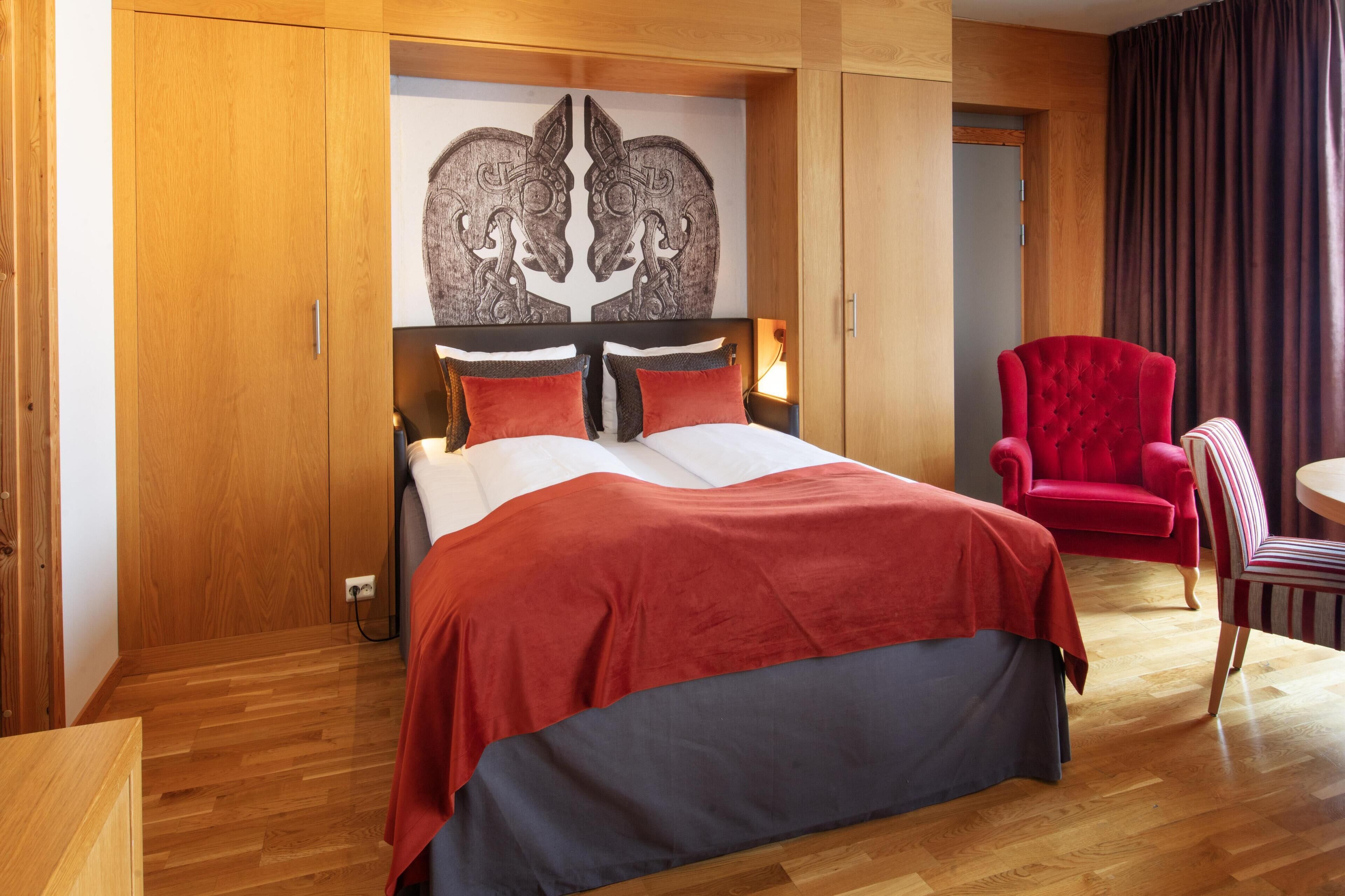 Una camera da letto con un letto, una sedia rossa, una sedia a righe, un armadio in legno e un quadro appeso al muro.