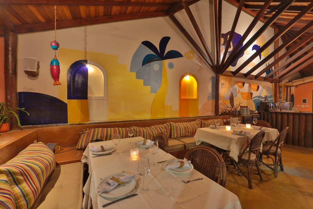 Un restaurant avec un plafond haut, des poutres apparentes et une fresque colorée sur le mur. On y trouve plusieurs tables, décorées de bougies, ainsi qu’un banc le long du mur.
