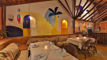 Un restaurant avec un plafond haut, des poutres apparentes et une fresque colorée sur le mur. On y trouve plusieurs tables, décorées de bougies, ainsi qu’un banc le long du mur.