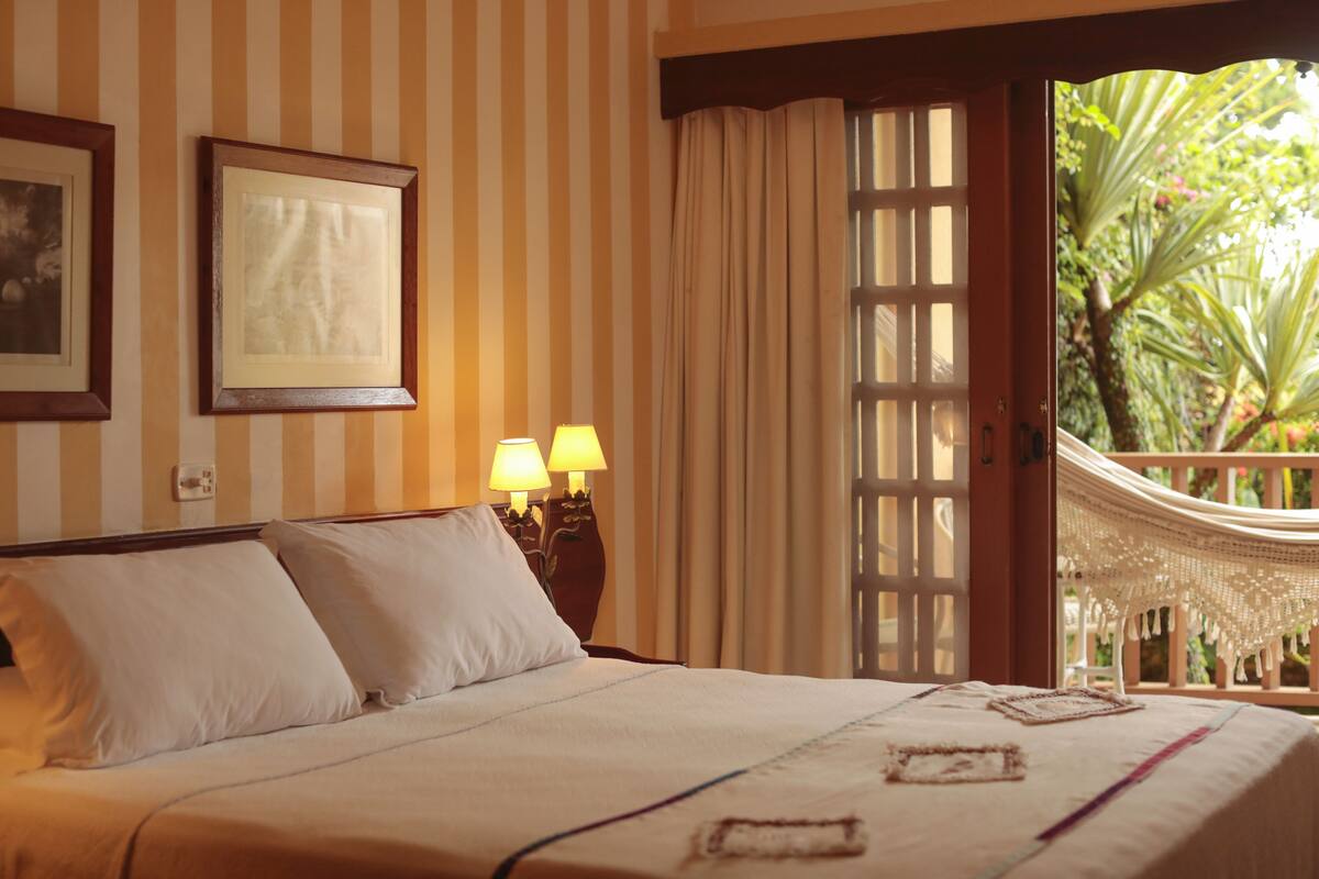 A hotel room with a large bed, striped wallpaper, two bedside lamps, a framed picture, and a view of a garden through a door.