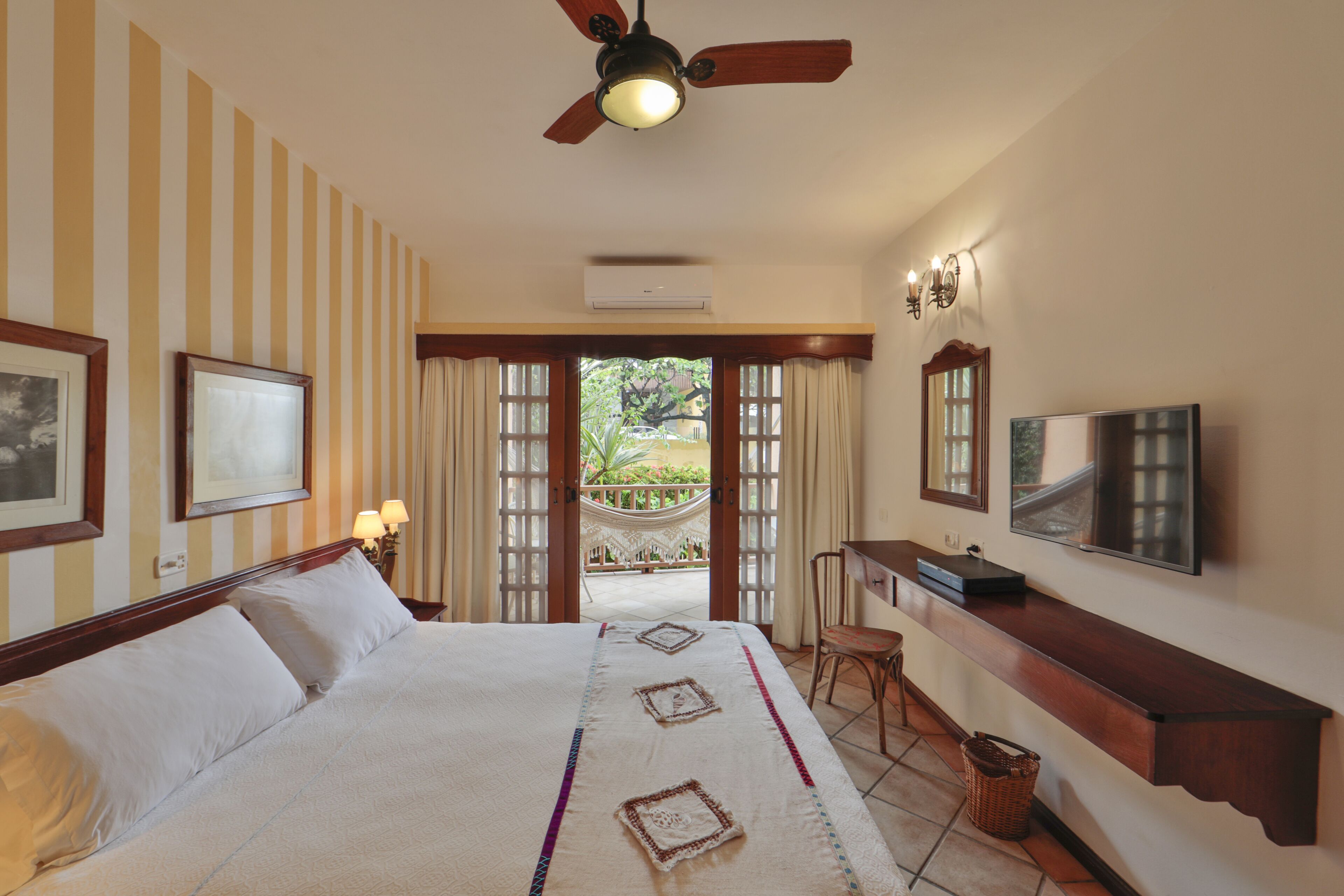 Habitación de hotel con cama, escritorio, silla, televisor y balcón con cortinas.