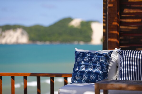Una terraza de madera con una cama adornada con almohadones azules y blancos, con vista a una playa de aguas turquesas cristalinas y colinas verdes al fondo.