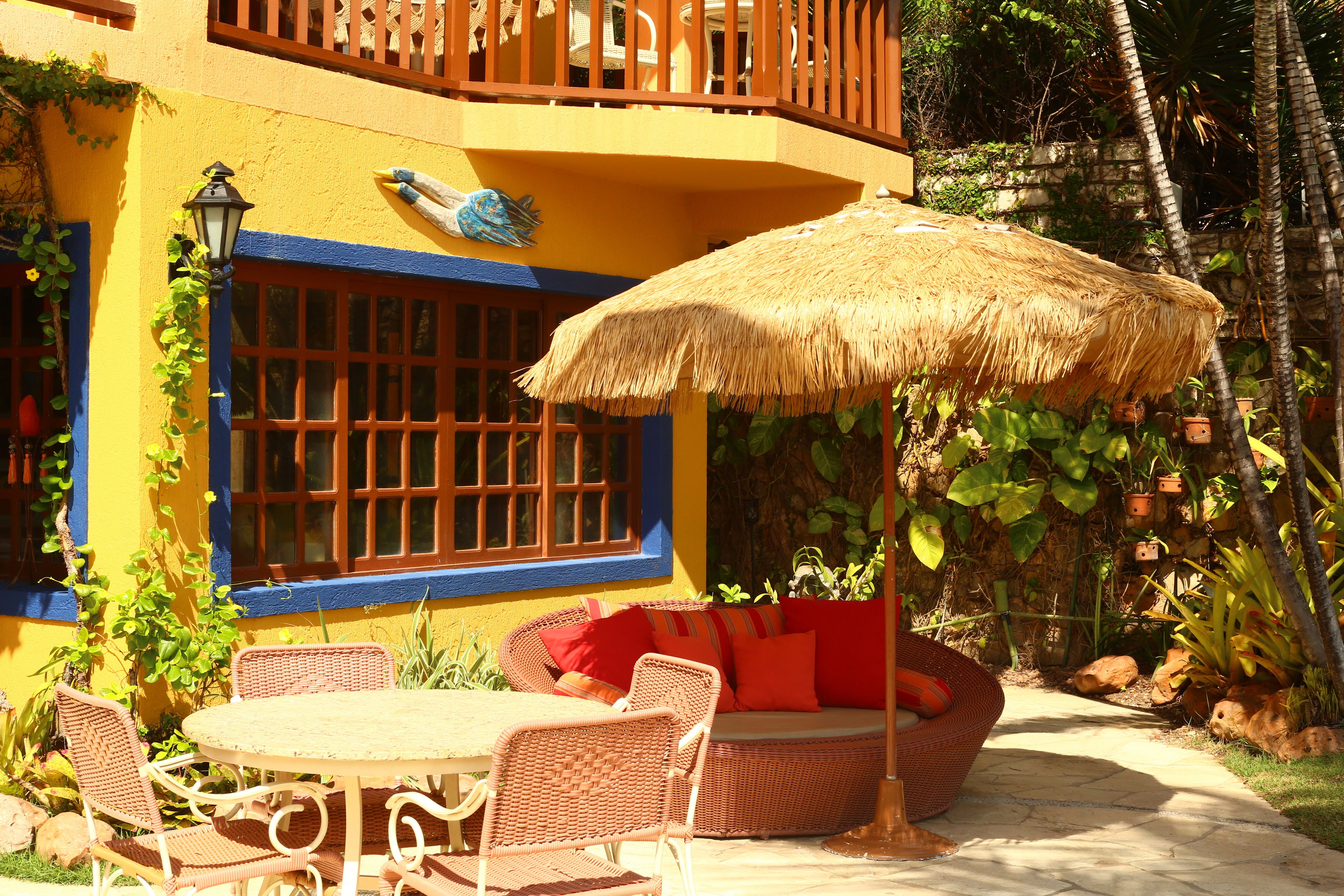 Un patio con mobiliario de mimbre, un paraguas de paja y vistas a un edificio amarillo con detalles azules y un balcón.