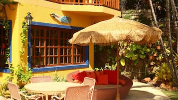Un patio con mobiliario de mimbre, un paraguas de paja y vistas a un edificio amarillo con detalles azules y un balcón.