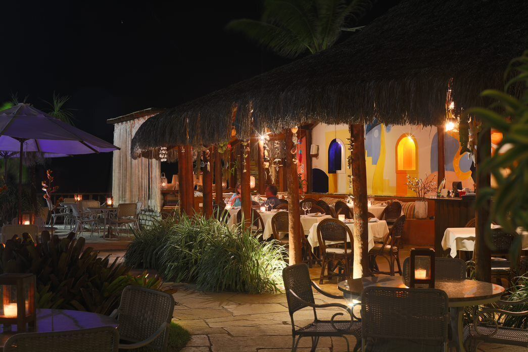 An outdoor dining area with thatched-roof structures, wicker furniture, and ambient lighting.