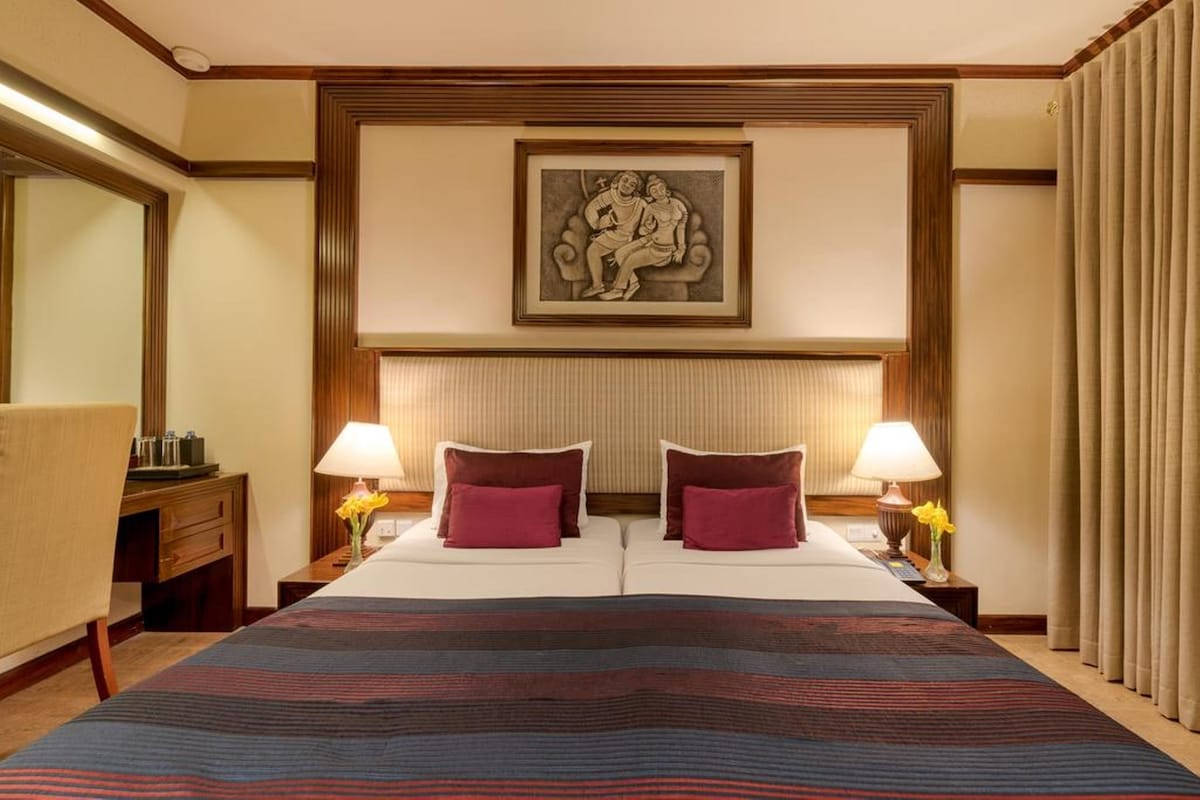 A hotel room with a large bed, two bedside lamps, a headboard with a framed artwork, a nightstand with a lamp, and a window with curtains.