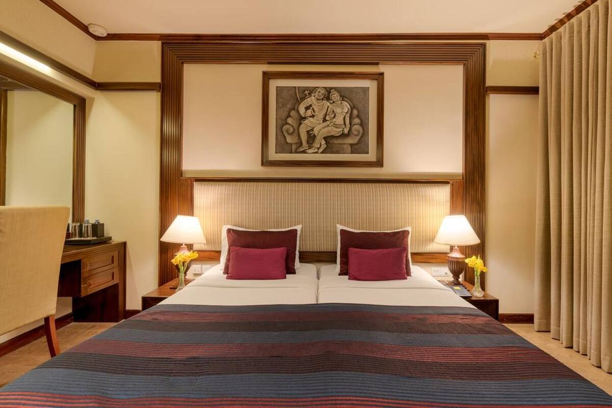 A hotel room with a large bed, two bedside lamps, a headboard with a framed artwork, a nightstand with a lamp, and a window with curtains.