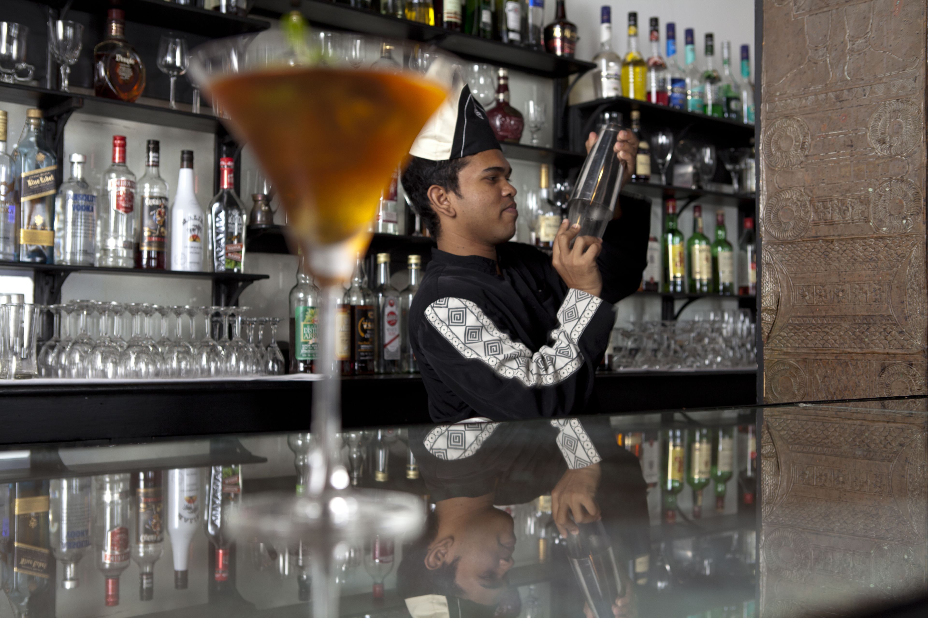 A bar with a bartender preparing a drink, shelves stocked with various bottles, and a glass of cocktail in the foreground.