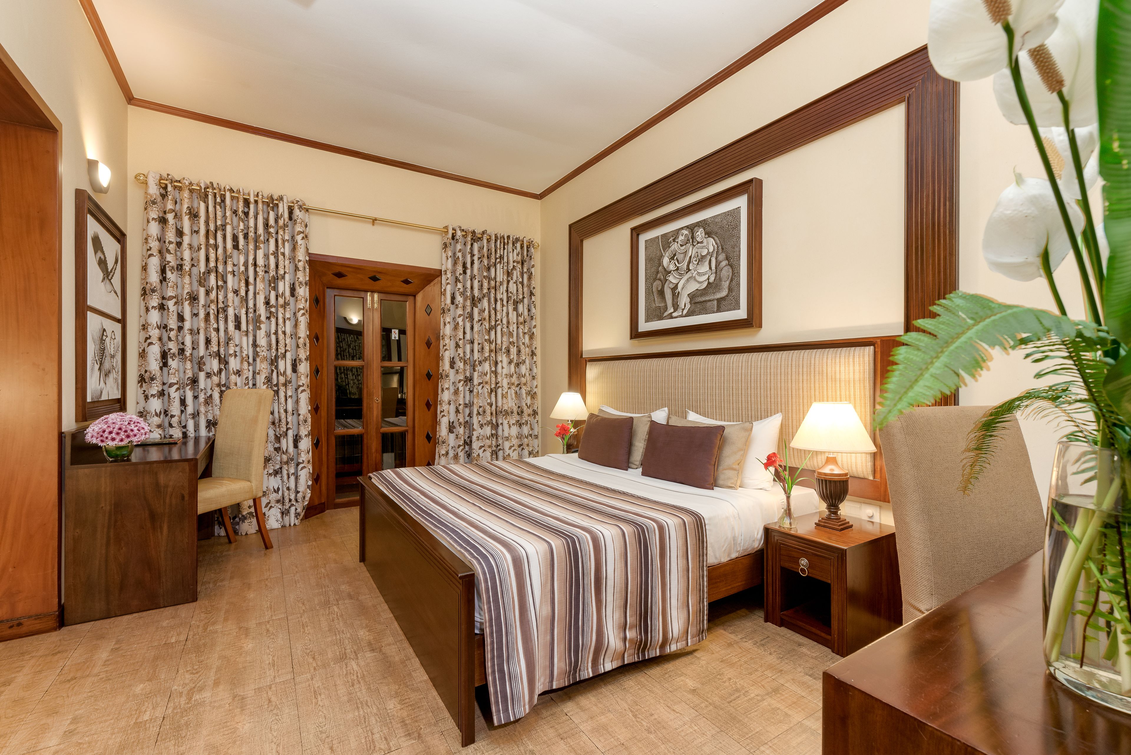 A hotel room with a large bed, two bedside tables, a desk, and a chair. There is a framed picture on the wall and a potted plant on the desk.
