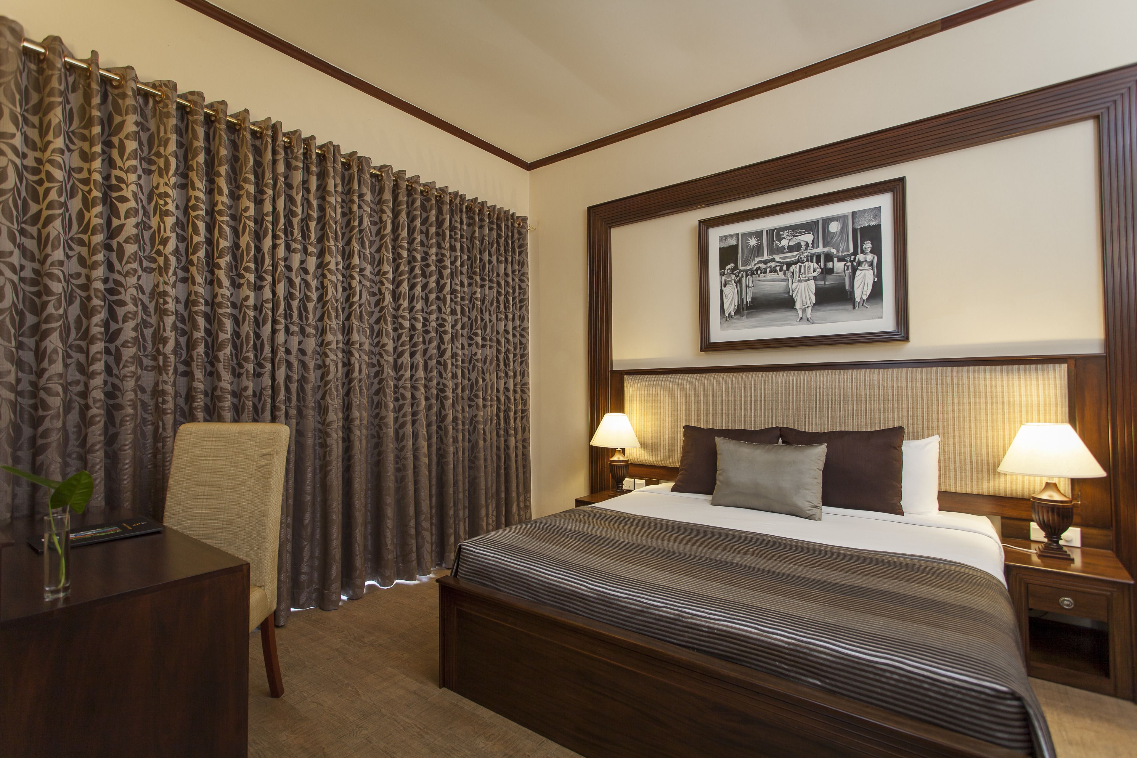 Una habitación de hotel con una cama grande, dos mesitas de noche con lámparas, una silla, un escritorio con una planta y una cortina con estampado de hojas.