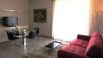 A living room with a red sofa, a glass coffee table, a dining table with chairs, a flat-screen TV, and a painting on the wall.