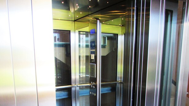 Interior d'un ascensor amb parets metàl·liques, una porta de vidre i un sòl amb un disseny de rajoles fosques.
