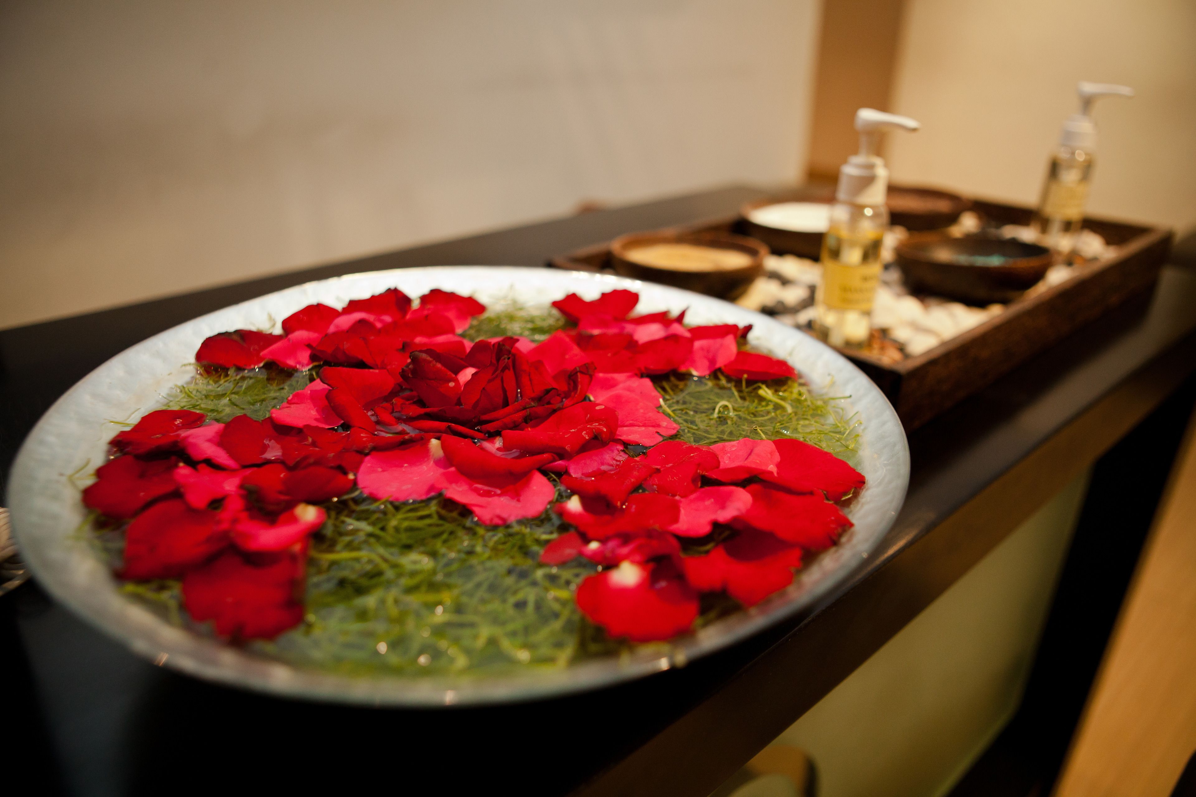 Un ambiente de spa con una bandeja decorativa que contiene pétalos de rosa y un cuenco de hojas verdes.