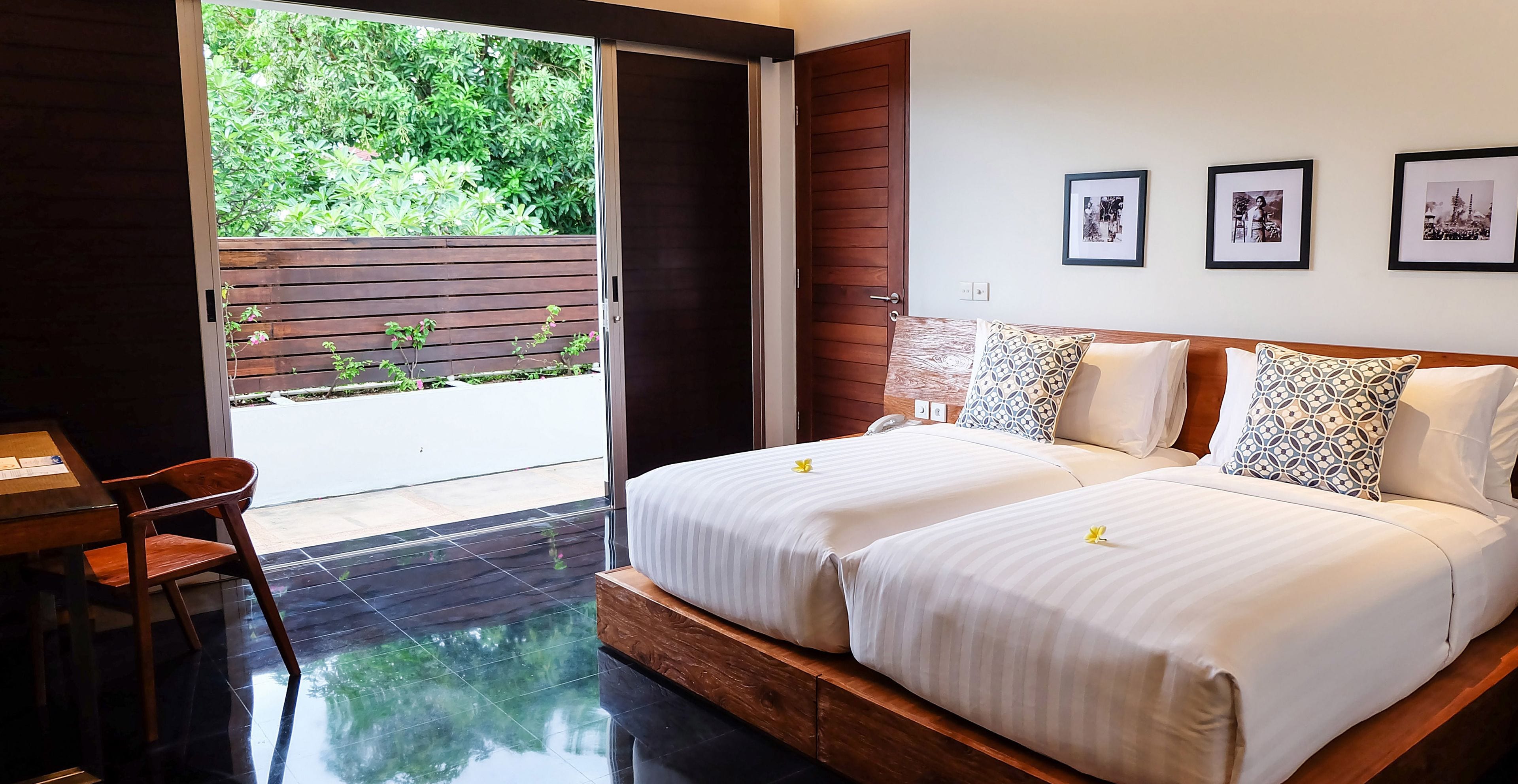 A bedroom with two beds, a wooden headboard, a desk with a chair, and a view of greenery through a sliding door.