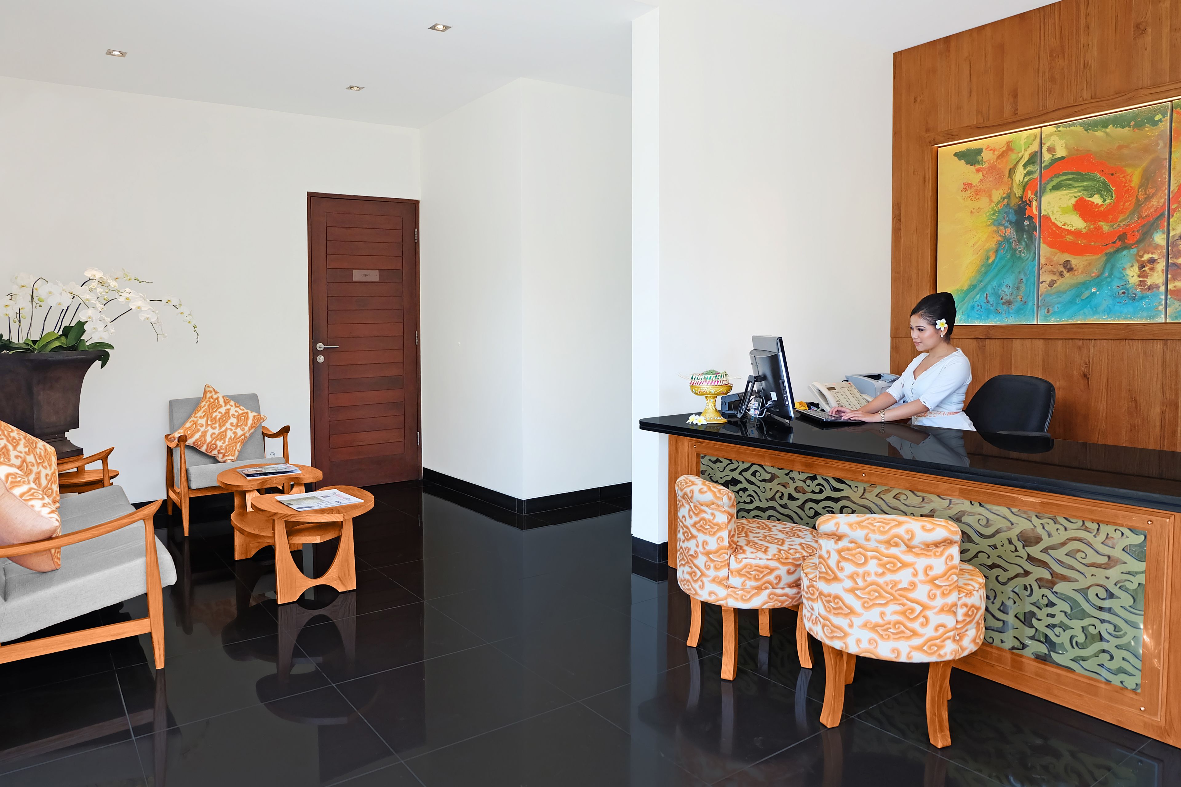 A reception area with a receptionist, a wooden reception desk, patterned chairs, a potted plant, and a colorful abstract painting on the wall.