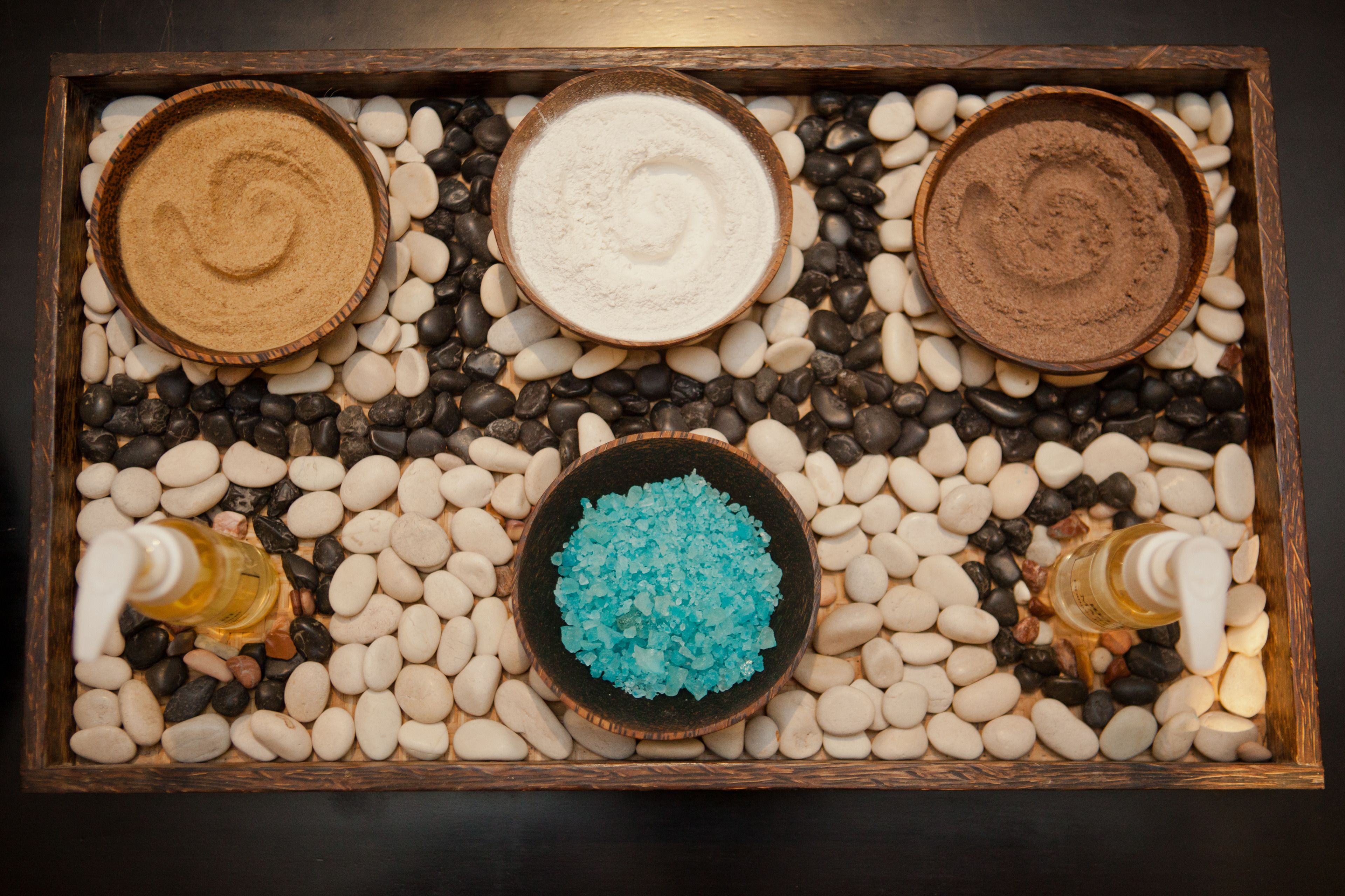 A tray with three bowls of different powders and a bowl of blue crystals, surrounded by white and black pebbles.