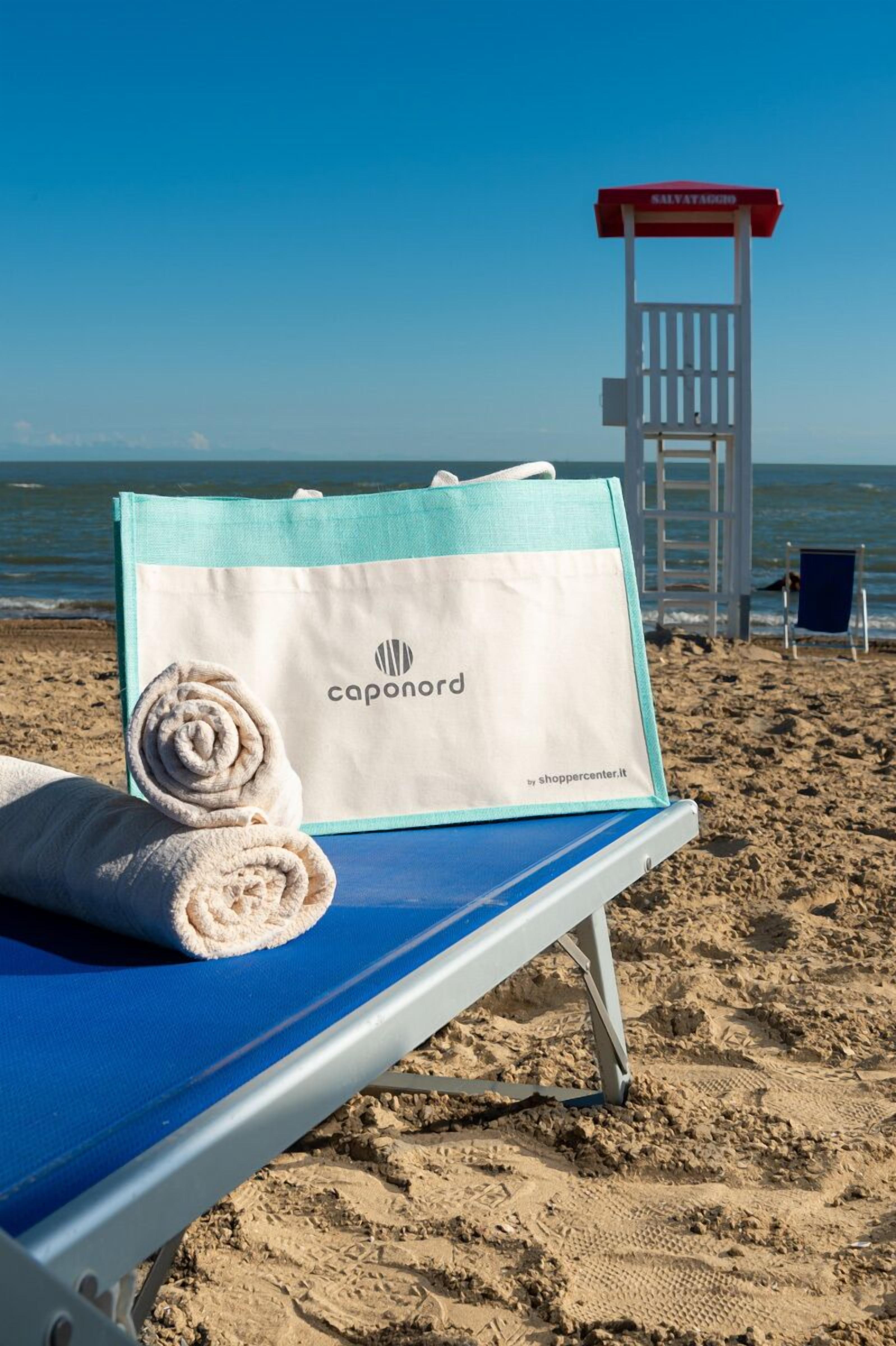 A beach chair with a towel and a shopping bag labeled "caponord" on a sandy beach.