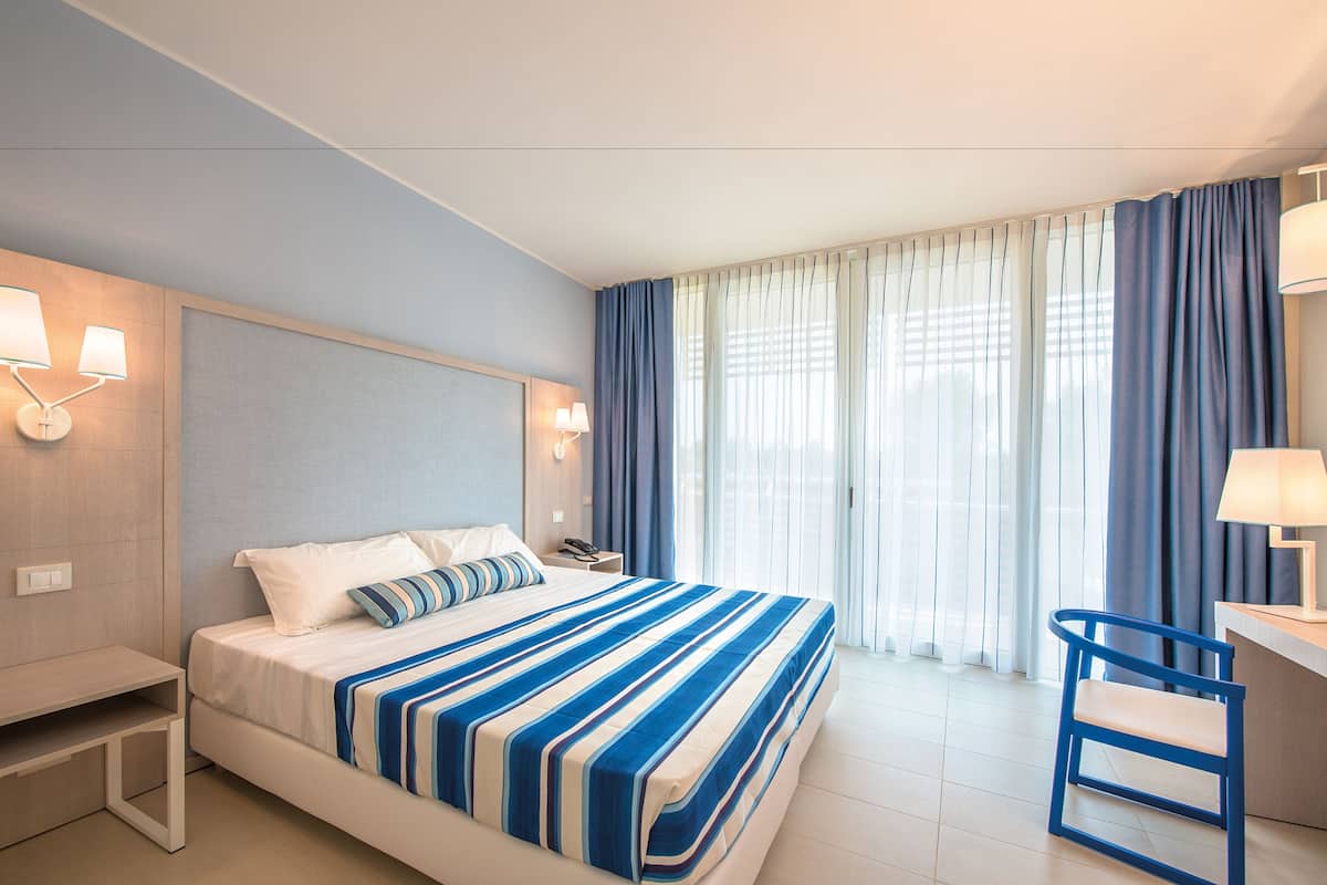 Quarto de hotel com cama, mesa de cabeceira, luminária, cadeira e cortinas.