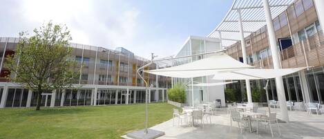 Un patio exterior moderno con mesas y sillas, rodeado por un edificio de vidrio con techo blanco.
