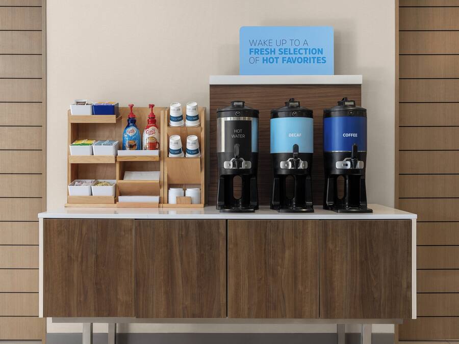 A coffee station with hot water, decaf, and coffee dispensers, and a sign above stating 'WAKE UP TO A FRESH SELECTION OF HOT FAVORITES.'