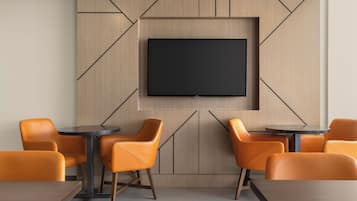 Una habitación moderna con una televisión de pantalla plana montada en una pared con diseño geométrico, rodeada de sillas y mesas naranjas.