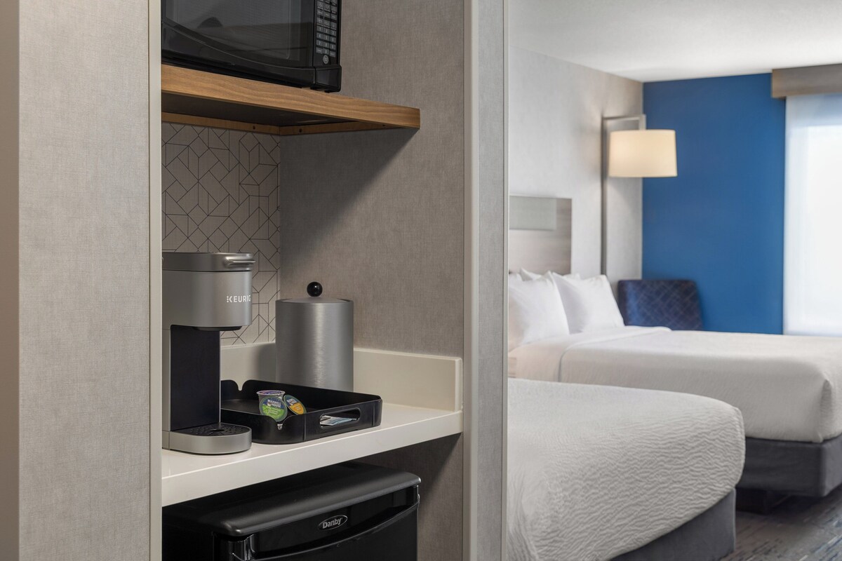 A hotel room with a kitchenette, a bed, a nightstand, and a wall-mounted lamp.