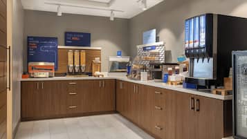 A modern kitchen area with wooden cabinets, a coffee machine, a bakery display, and a beverage dispenser.