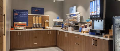 A modern kitchen area with wooden cabinets, a coffee machine, a bakery display, and a beverage dispenser.