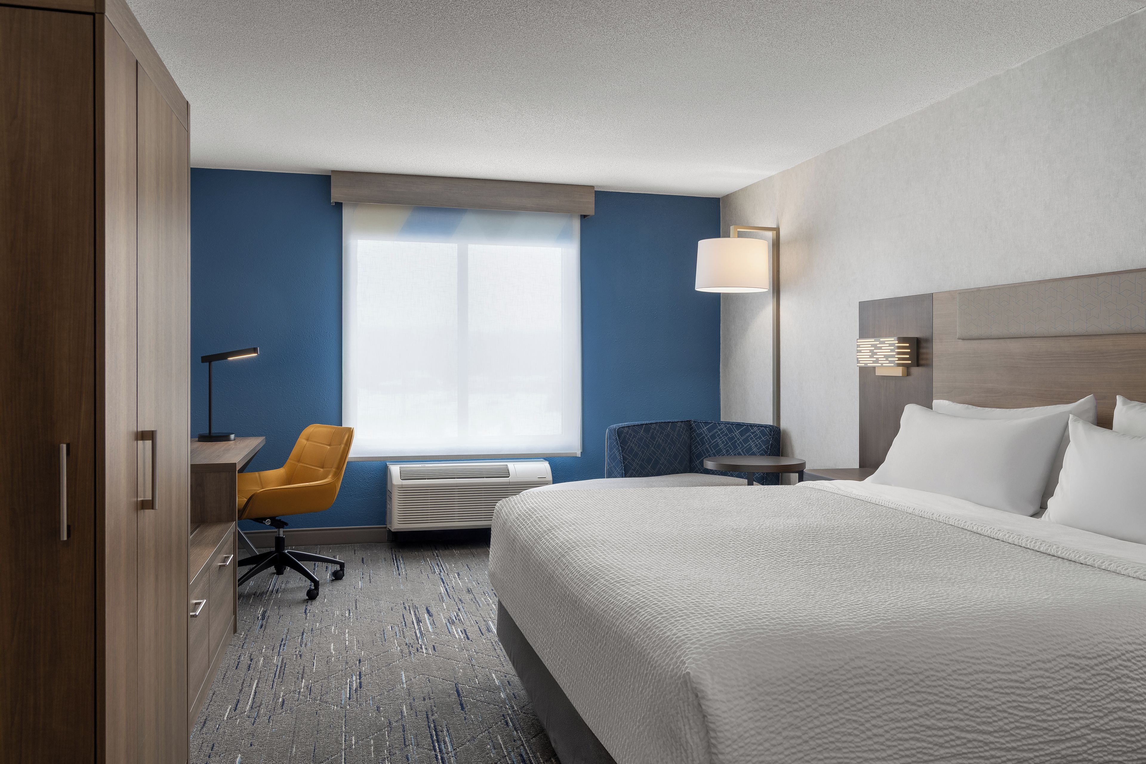 A hotel room with a large bed, a desk with a chair, a window with curtains, and a wall-mounted lamp.