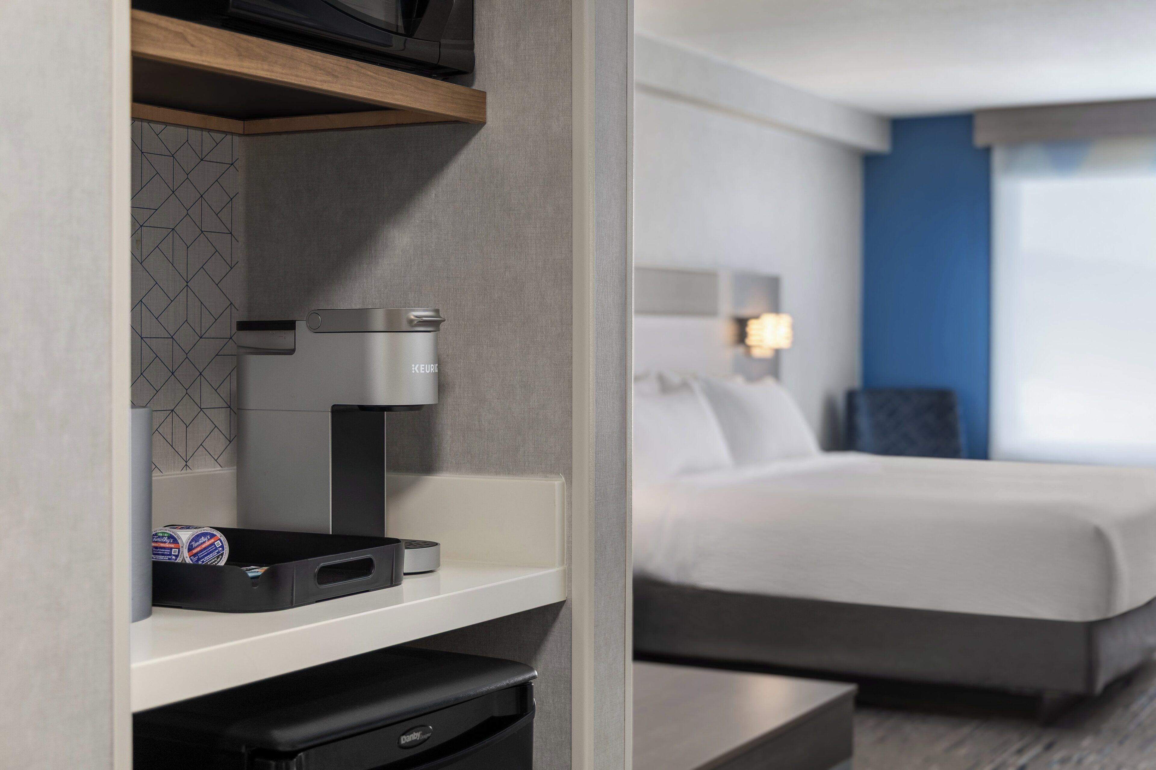 Une chambre d’hôtel équipée d’un lit, d’une machine à café, d’une poubelle et d’une étagère sur laquelle se trouvent une boîte de conserve et une boîte.
