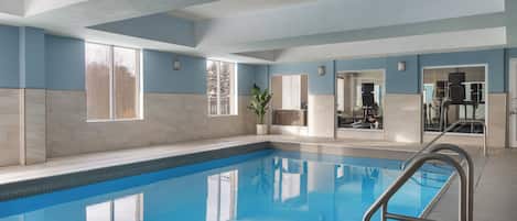 An indoor swimming pool with clear blue water, a metal ladder, and a view of the outside through large windows.