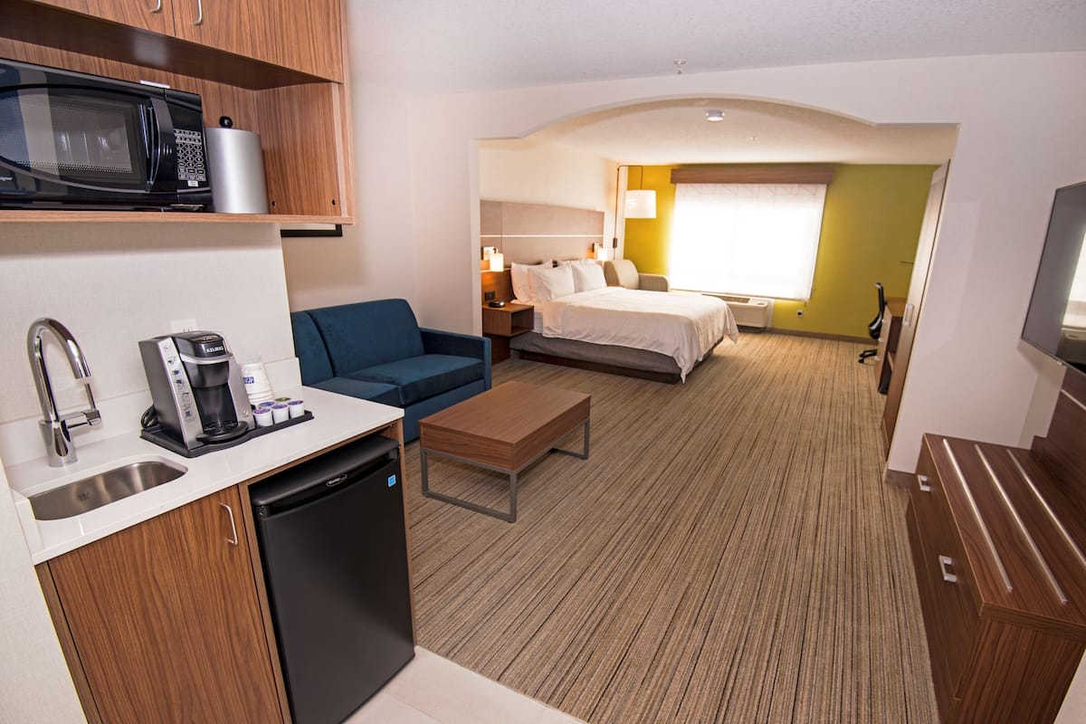 A compact hotel room with a kitchenette, a bed, a sofa, and a television.
