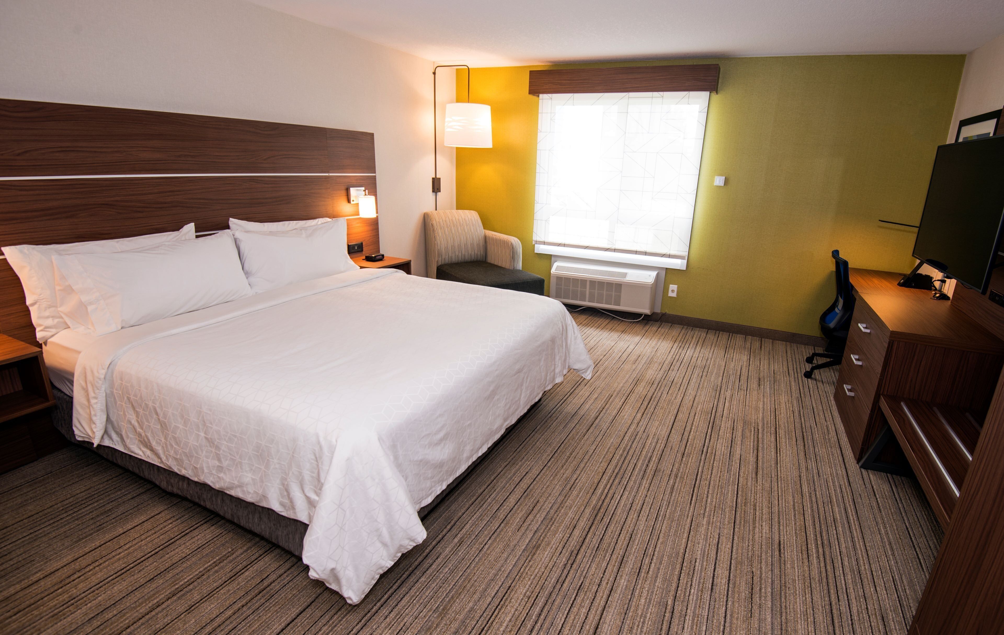 Une chambre d’hôtel avec un grand lit, une chaise, un bureau avec une télévision et une fenêtre avec des stores.