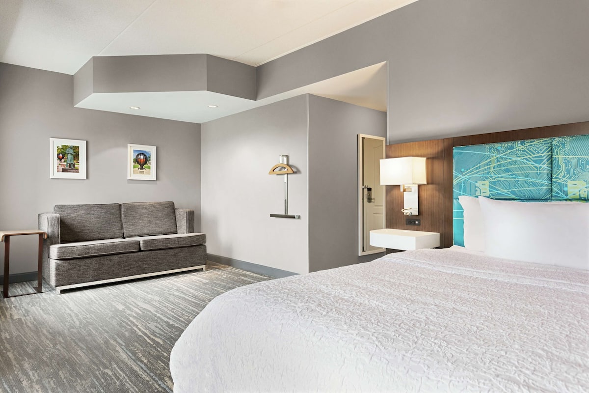 Ein Hotelzimmer mit einem großen Bett, einer Sitzecke, einem Schreibtisch, einer Lampe und einem Badezimmer mit Spiegel und Waschbecken.