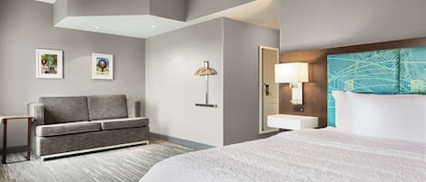 A hotel room with a large bed, a sofa, a desk, a lamp, and a bathroom with a mirror and sink.