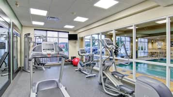 A gym with treadmills, elliptical machines, and a stationary bike, with a pool visible through the windows.