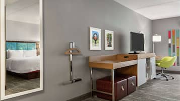 Una habitación de hotel con cama, escritorio, silla, televisor, espejo y obras de arte en la pared.