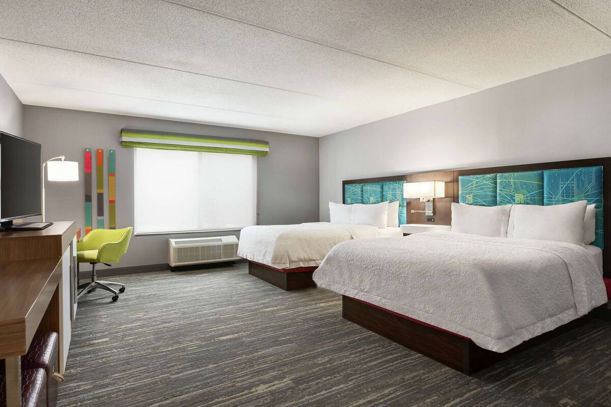 Ein Hotelzimmer mit zwei Betten, einem Schreibtisch mit Computer, einem Stuhl und einem Fenster mit Jalousien.