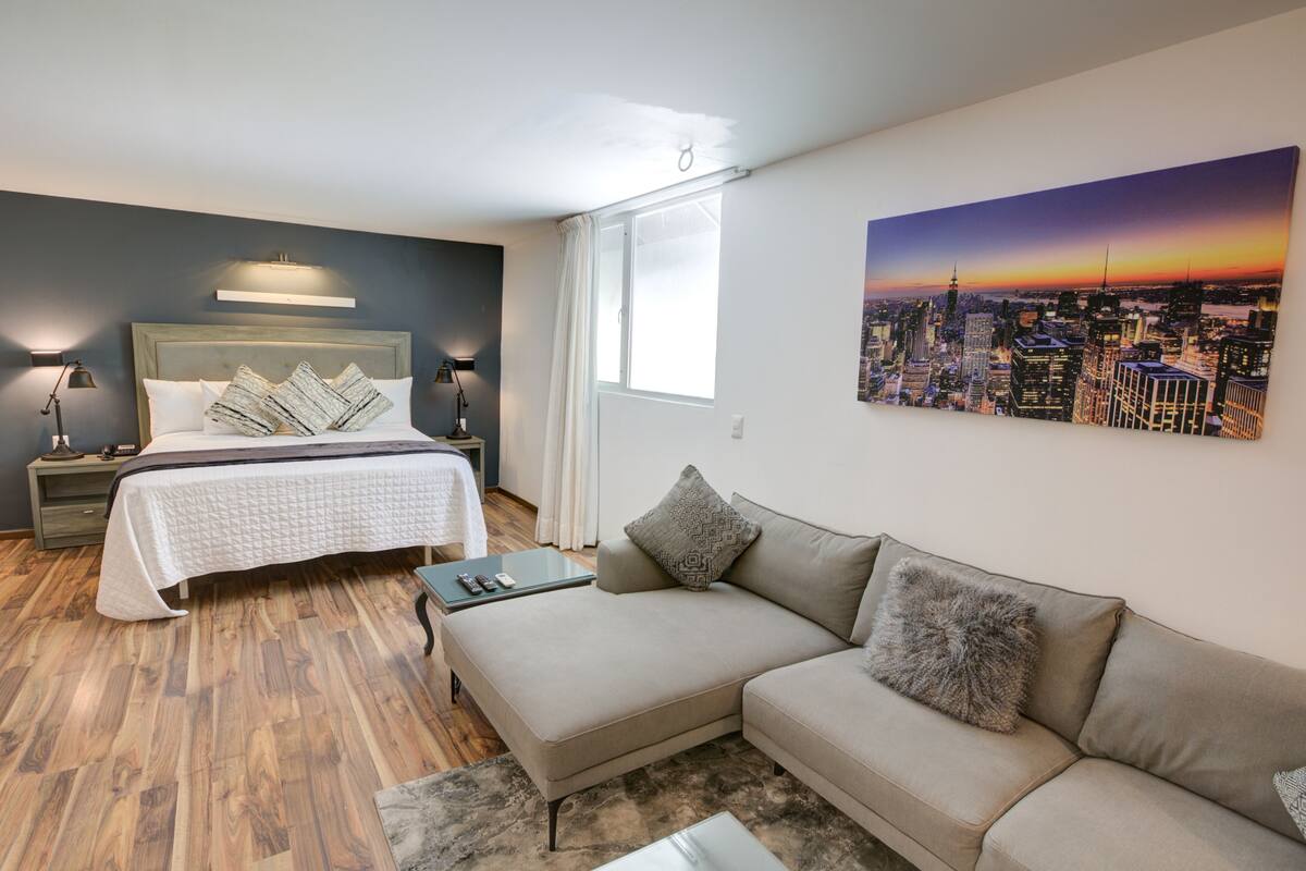 A modern hotel room with a bed, a sofa, a cityscape painting, and a window with curtains.